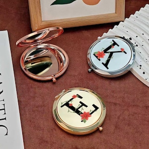 May include: Three compact mirrors. One is open, showing the mirror surface, with a rose gold finish. Two round compacts feature a letter and floral design. One has a silver finish, the other gold. All are on a brown surface.