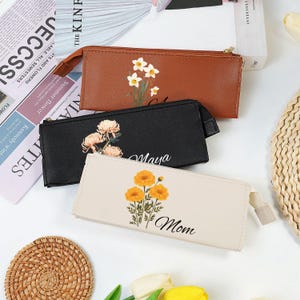 Op de afbeelding: Drie rechthoekige etuis met ritssluiting in bruin, zwart en crème. Elke etui heeft een bloemenontwerp en een naam: "A", "Maya" en "Mom". De etuis liggen op een wit oppervlak met een boek en een geweven onderzetter.