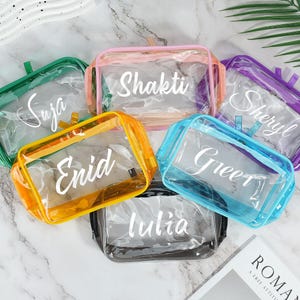 May include: A collection of clear, rectangular cosmetic bags in various colors, including green, pink, yellow, blue, and purple. Each bag has a white name printed on it. The bags are arranged in a circular pattern.
