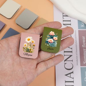 May include: Two pink and green leather magnetic bookmarks with floral designs. The pink bookmark has a daisy design and the text "Megan". The green bookmark has a water lily design and the text "Lula".