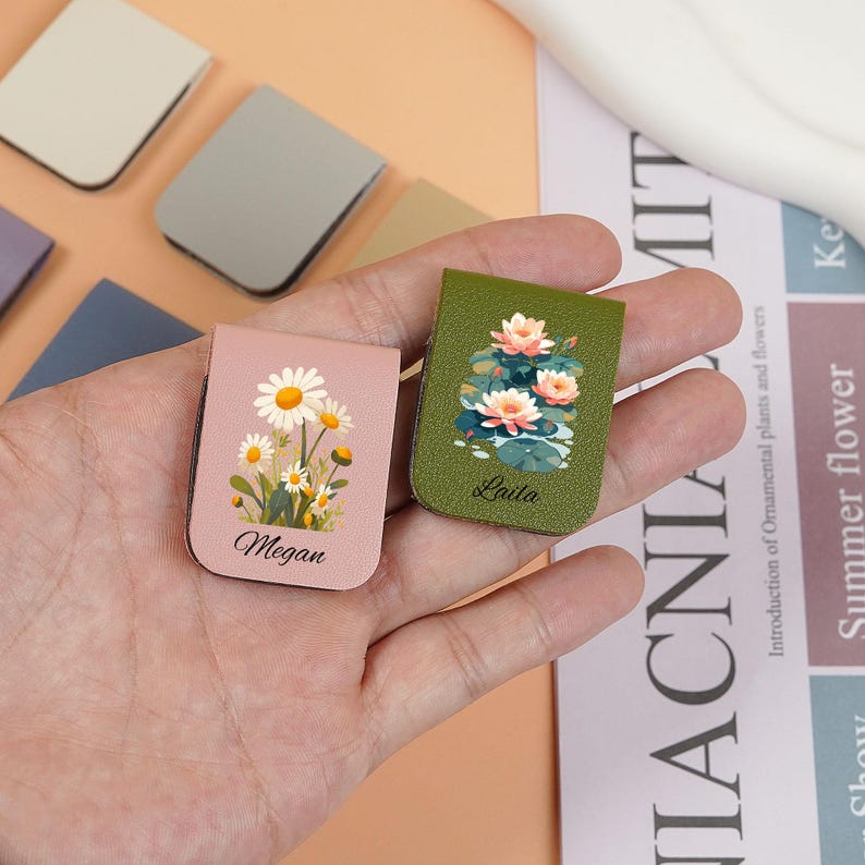 May include: Two pink and green leather magnetic bookmarks with floral designs. The pink bookmark has a daisy design and the text "Megan". The green bookmark has a water lily design and the text "Lula".