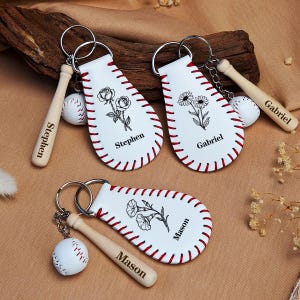 Könnte beinhalten: Drei weiße, baseballförmige Schlüsselanhänger aus kunstlederartigem Material mit roten Nähten, jeweils mit einer schwarzen Blumenillustration und einem Namen: Stephen, Gabriel oder Mason. Jeder Anhänger enthält einen kleinen Holzschläger.