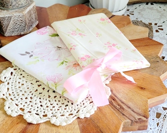 May include: Two fabric-covered journals with a pink ribbon tied around them. The journals have a floral pattern with pink and white flowers on a cream background.