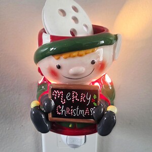May include: A ceramic elf-shaped plug-in wax warmer featuring a green hat, red clothing, and black gloves. The elf holds a small rectangular sign with the text Merry Christmas in red and white candy cane lettering against a black background.