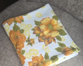 May include: Folded white towel with a floral pattern in shades of orange, yellow, and green. The flowers are large and stylized, set against a white background. The towel is folded neatly, suggesting it's ready for use.
