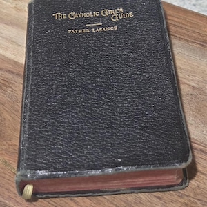May include: A small, black book titled "The Catholic Girl's Guide" by Father Lasance. The book has gold lettering and a gold ribbon bookmark. The book is resting on a wooden surface.