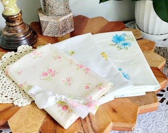 May include: Four fabric squares with floral patterns. The squares are folded and stacked on top of each other. The top square has a blue floral pattern, the second square has a yellow floral pattern, the third square has a pink floral pattern, and the bottom square has a white floral pattern.