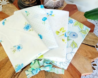 May include: Four white fabric squares with blue and green floral patterns. The fabric squares are arranged on a wooden surface.
