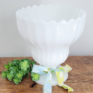 May include: White milk glass vase with a scalloped rim and fluted design. The vase is decorated with a blue and green floral ribbon.