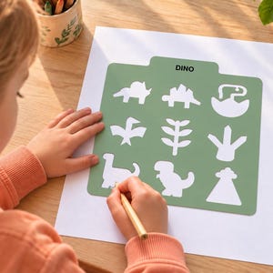 May include: A square sage green plastic stencil sheet featuring nine cut-out shapes of various dinosaurs and prehistoric elements, with the word DINO printed in black at the top center, positioned on a white sheet of paper for drawing.