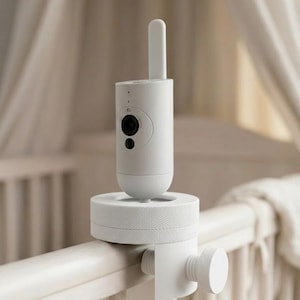May include: A white baby monitor camera with a cylindrical body and a rounded antenna. The camera is mounted on a white clamp, attached to a cot rail. The camera has a lens and a small button.