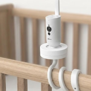 May include: A white baby monitor with a cylindrical body, a camera lens, and an antenna. The monitor is mounted on a white base and attached to a cot rail with white hooks. The brand name "Alecto" is printed on the monitor.