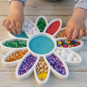 Op de afbeelding: Een witte, bloemvormige knutselorganizer gevuld met diverse kleurrijke items. De organizer bevat blauw zand, groene knikkers, rood zand, houten kralen, kleurrijke knopen, witte pompons, oranje kralen en paarse pailletten. De handen van een kind reiken in de organizer.