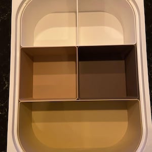 Puede incluir: Una bandeja de almacenamiento rectangular de plástico blanco con cinco compartimentos distintos. La sección superior tiene dos divisores blancos, la sección central contiene dos recipientes cuadrados en color beige y marrón oscuro, y la sección inferior es un gran recipiente beige.