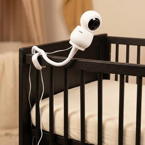Puede incluir: Una cámara de vigilancia para bebés blanca está sujeta a una cuna marrón oscuro. La cámara tiene un brazo flexible y está colocada para ver el interior de la cuna. La cuna tiene un colchón blanco y barras verticales.