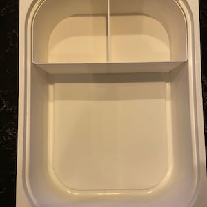 Pode incluir: Um recipiente branco de plástico para armazenamento de alimentos com uma seção superior dividida e um compartimento inferior maior. O recipiente tem cantos arredondados e uma superfície lisa, adequado para armazenar alimentos.