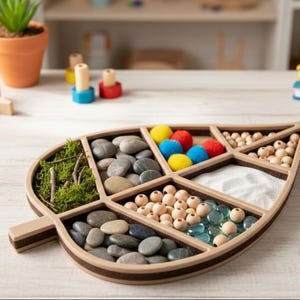 Puede incluir: Una bandeja sensorial de madera con forma de hoja con múltiples compartimentos llenos de diversas texturas. La bandeja contiene piedras lisas, pompones de colores, cuentas de madera, arena y cuentas de vidrio. Una pequeña maceta de terracota con una planta está en el fondo.