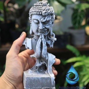May include: A gray statue depicting a split face, one side serene and the other demonic. The statue's hands are clasped in prayer, and it rests on a square base. The sculpture is held in a hand against a blurred background of greenery.
