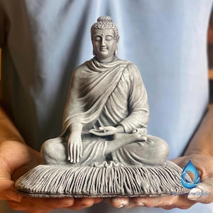 Puede incluir: Una estatua gris de Buda en posición de meditación sentado. La estatua está sobre una base texturizada y tiene detalles intrincados en las túnicas y los rasgos faciales. La estatua está sostenida en las manos de alguien.