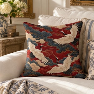 May include: A square throw pillow featuring a woven tapestry design of three white cranes with red crowns and black wing tips, flying amidst stylized dark blue and cream-colored clouds against a deep red background.