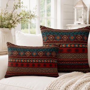 Op de afbeelding: Twee decoratieve kussens met een donkerrode basis en ingewikkelde horizontale banden van geometrische patronen in zwart, oranje en talingblauw. Het ene kussen is vierkant en het andere rechthoekig, beide met identieke herhalende motieven.