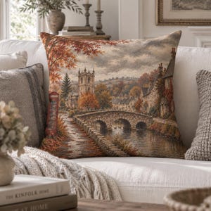 Könnte beinhalten: Ein quadratisches Zierkissen mit einem gewebten Wandteppichmotiv einer ländlichen englischen Dorfszene. Das Bild zeigt eine Steinbrücke über einen Fluss, einen historischen Kirchturm, herbstliches Laub in Orange- und Brauntönen sowie eine rote Telefonzelle.