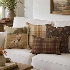May include: A collection of four decorative throw pillows arranged on a white sofa. One square pillow features a woven scene of two hunting dogs in a field, one is a tan plaid, one is a dark brown floral print, and one is a rectangular brown plaid.