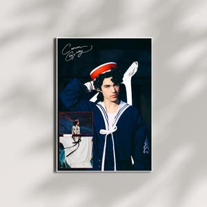 Könnte beinhalten: Ein gerahmtes Poster mit einer Person in einem marineblauen Outfit im Matrosenstil mit weißem Kragen und rot-weißem Hut. Das Poster enthält den handgeschriebenen Namen "Conan Gray" in der oberen linken Ecke und zwei kleinere inset-Bilder.