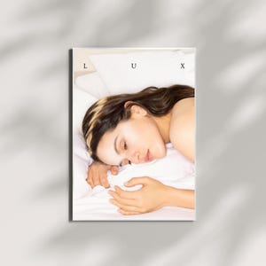 May include: A rectangular print featuring a person lying on a white pillow and sheets. The person has long brown hair and is resting their head on their arm. The word "LUX" is printed in capital letters at the top of the print.