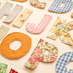 May include: Assortment of colorful, fabric-covered letters in various patterns, including gingham, floral, and solid colors. The letters are arranged on a light-colored surface, creating a decorative and playful display. Ideal for crafting or home decor.