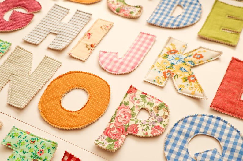 May include: Assortment of colorful, fabric-covered letters in various patterns, including gingham, floral, and solid colors. The letters are arranged on a light-colored surface, creating a decorative and playful display. Ideal for crafting or home decor.
