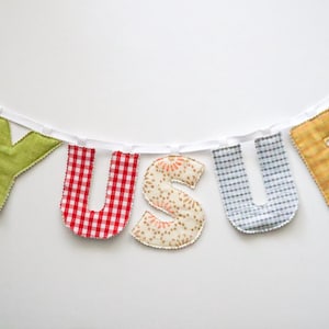 May include: A handmade banner spelling "YUSUF" with fabric letters. The letters are in various patterns and colors, including green, red gingham, floral, blue gingham, and gold. The letters are strung on a white ribbon.