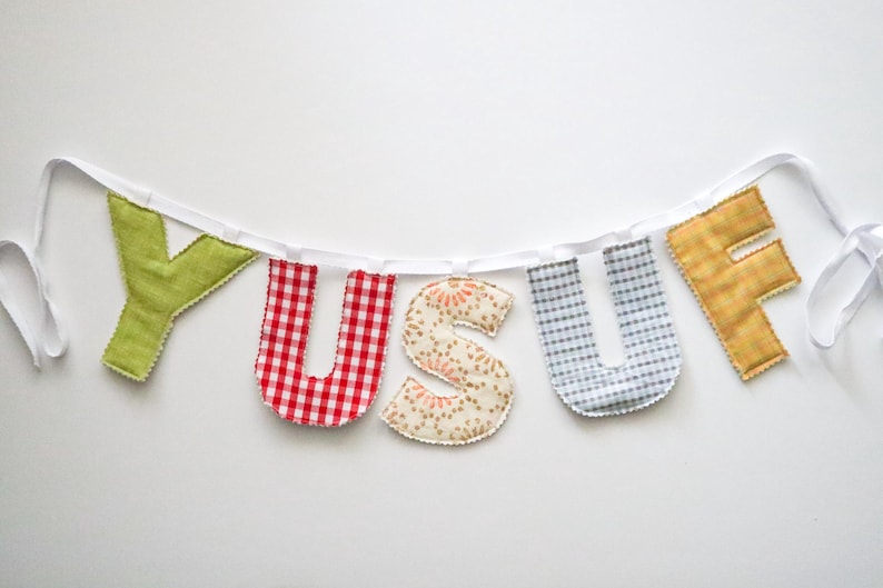 May include: A handmade banner spelling "YUSUF" with fabric letters. The letters are in various patterns and colors, including green, red gingham, floral, blue gingham, and gold. The letters are strung on a white ribbon.