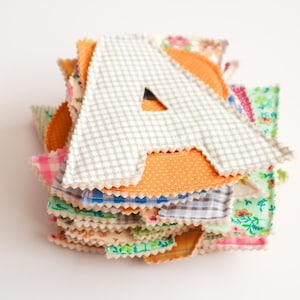 May include: A stack of colorful fabric letters, with the letter "A" prominently displayed. The "A" is made of a light gray and white checkered fabric. Other fabrics include orange polka dots, floral prints, and pink gingham.