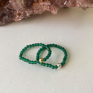 May include: Two green beaded rings with a gold and silver accent bead. The rings are set against a backdrop of a large, dark purple crystal cluster and a white surface.