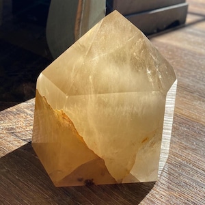 May include: A large, clear, light brown crystal in the shape of a pyramid. The crystal is sitting on a wooden surface.