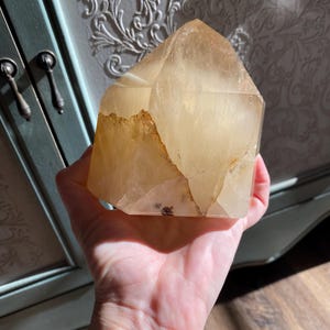 Puede incluir: Una gran punta de cristal de citrino dorado, transparente, con una textura natural, cruda y rugosa. El cristal se sostiene en una mano, mostrando su tama&ntilde;o y forma.