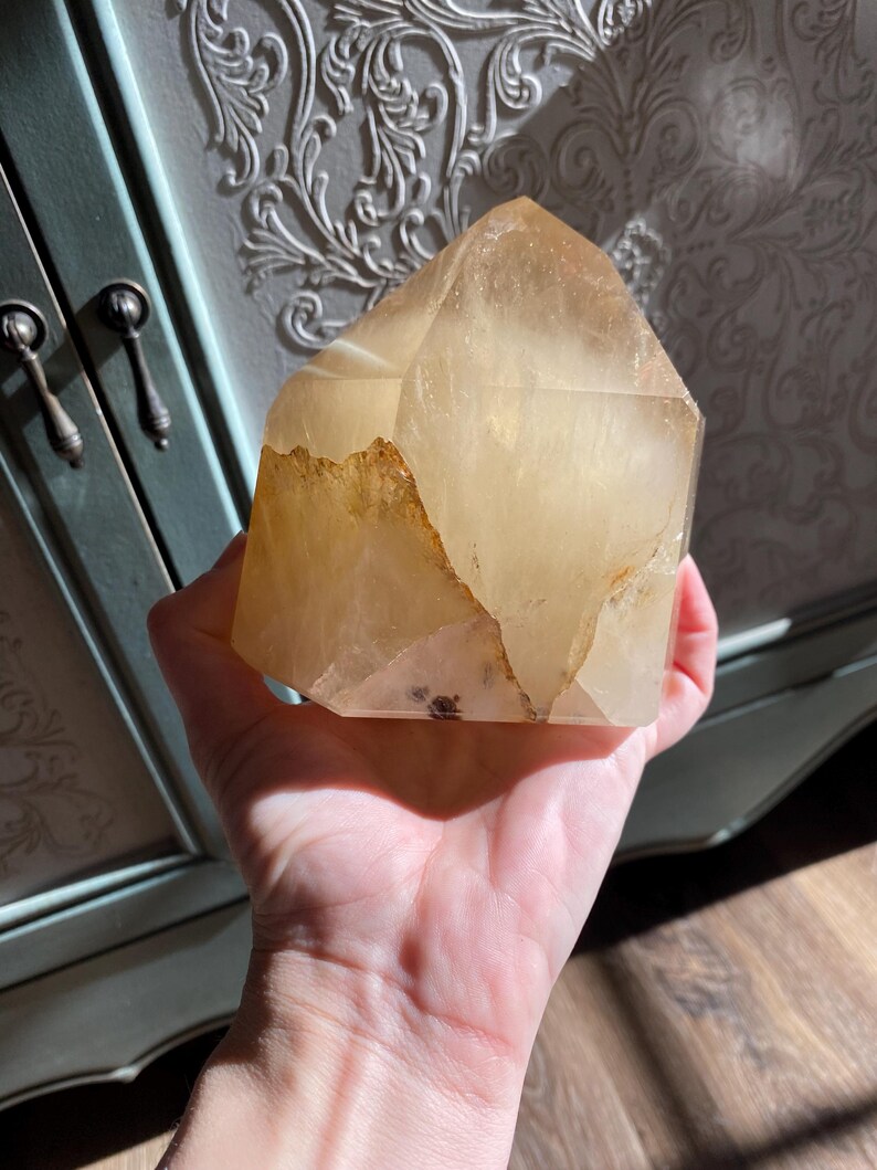 Puede incluir: Una gran punta de cristal de citrino dorado, transparente, con una textura natural, cruda y rugosa. El cristal se sostiene en una mano, mostrando su tama&ntilde;o y forma.
