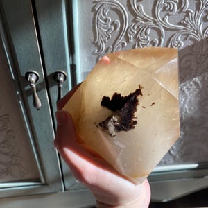 Puede incluir: Un cristal claro, facetado, de color marr&oacute;n claro con una inclusi&oacute;n marr&oacute;n oscura. El cristal tiene forma de pir&aacute;mide y se sostiene en una mano.