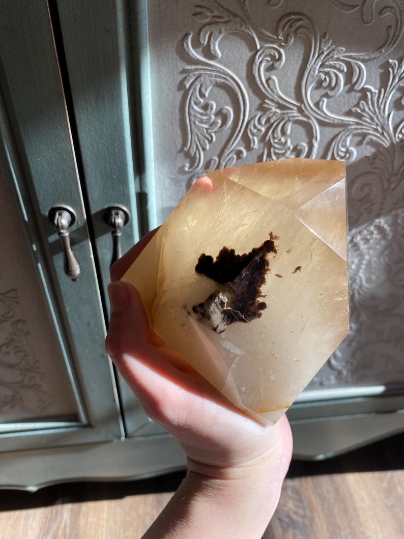 Puede incluir: Un cristal claro, facetado, de color marr&oacute;n claro con una inclusi&oacute;n marr&oacute;n oscura. El cristal tiene forma de pir&aacute;mide y se sostiene en una mano.