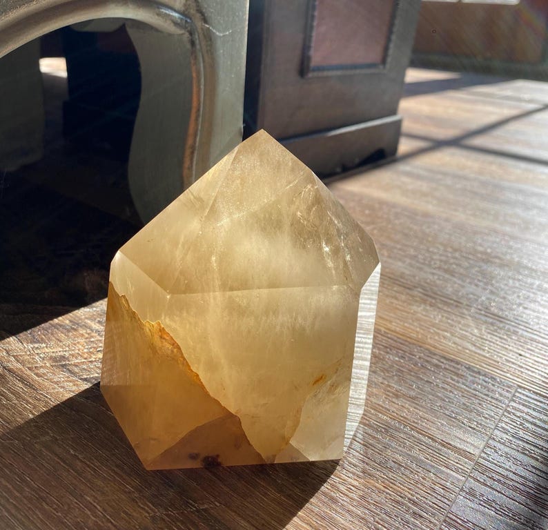 May include: A large, clear, light brown crystal in the shape of a pyramid. The crystal is sitting on a wooden surface.
