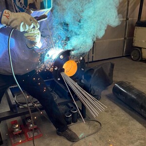 May include: A welder working on a metal pipe with a welding torch. The pipe is attached to a metal stand and there are several metal rods on the stand. The welder is wearing a welding mask and gloves. There is blue smoke coming from the welding torch.