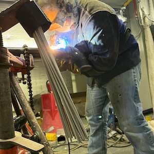 May include: A person welding metal rods with a bright blue flame, wearing a welding helmet, gloves, and a dark blue jacket. The rods are held in a black clamp, and the scene is illuminated by the welding arc.