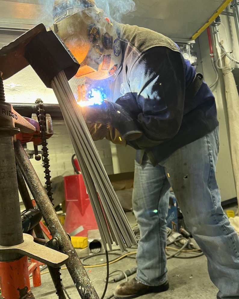 May include: A person welding metal rods with a bright blue flame, wearing a welding helmet, gloves, and a dark blue jacket. The rods are held in a black clamp, and the scene is illuminated by the welding arc.