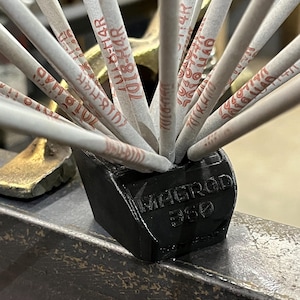 May include: A black 3D-printed holder labeled "MAGROD 360" contains multiple welding rods. The rods are light gray with red text. The holder sits on a metal surface, with a gold-colored object in the background.