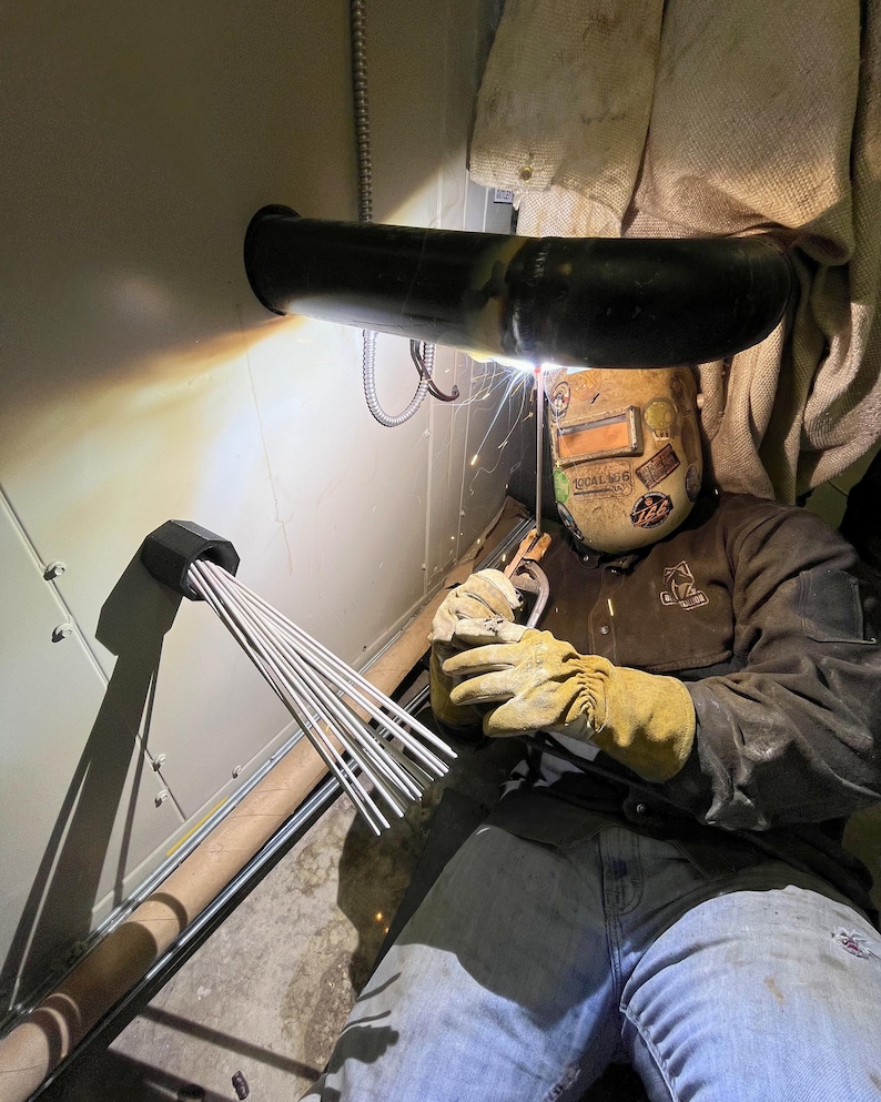 May include: A welder in a protective helmet and gloves is working on a metal pipe. Sparks fly as the welding torch is used. A bundle of welding rods is visible, along with a section of pipe and a wall.