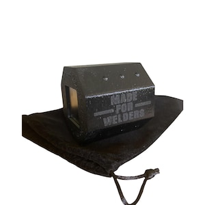 May include: A black metal welding tool with the text "MADE FOR WELDERS" engraved on its side. The tool is resting on a black drawstring bag against a white background. The tool has a rectangular shape with a small window.