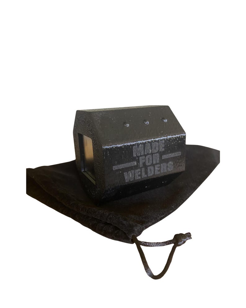 May include: A black metal welding tool with the text "MADE FOR WELDERS" engraved on its side. The tool is resting on a black drawstring bag against a white background. The tool has a rectangular shape with a small window.