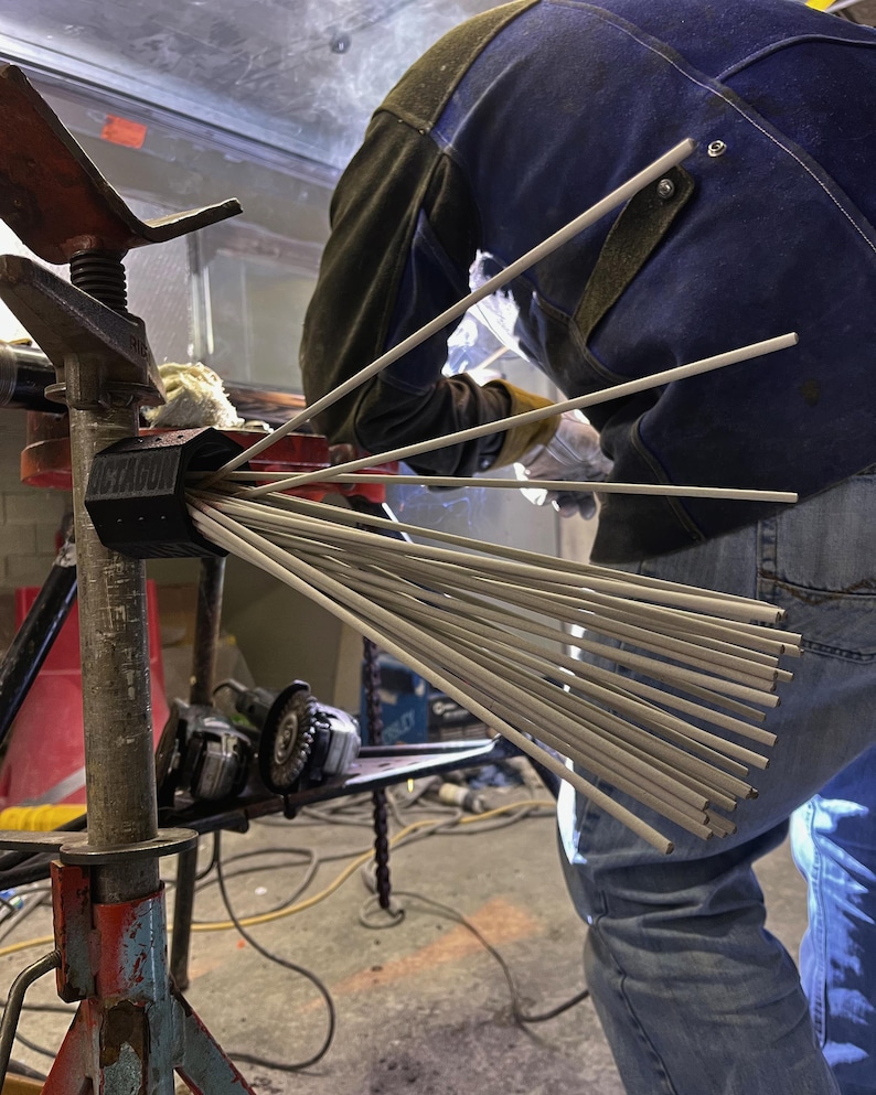 May include: A bundle of welding rods fanned out from a black container labeled "OCTAGON". A person in a blue denim jacket and welding gloves is working on a metal project. A grinder and other tools are visible in the background.