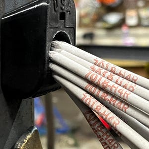 May include: Close-up of a black welding rod holder with the text "USA" embossed on the top. Several gray welding rods are inserted into the holder, each marked with "7018-H4R" in red ink. The rods are bundled together.